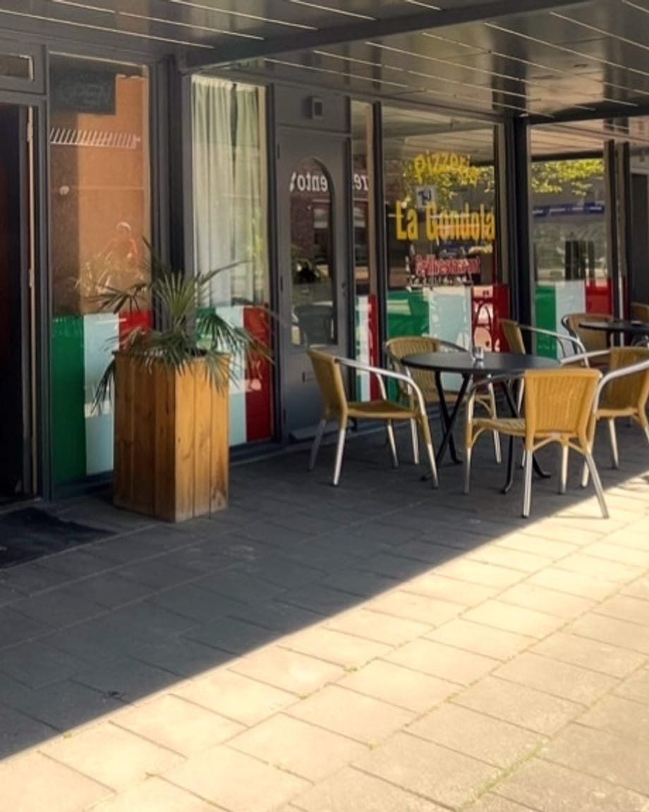 Het terras van La Gondola onder de overkapping, met rieten stoelen en ronde tafels